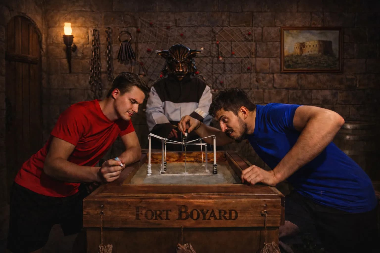 Двое игроков склонились над деревянным столом с надписью Fort Boyard и проходят испытание, за ними стоит персонаж в маске